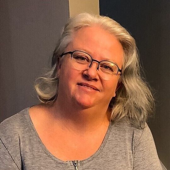 A woman with shoulder-length gray hair and glasses is smiling softly. She is wearing a light gray top and is in front of a neutral background.