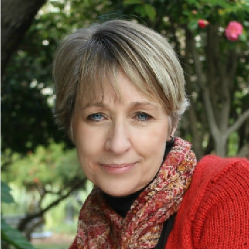 A middle-aged woman with short blonde hair smiles softly at the camera. She is wearing a red jacket and a patterned scarf. The background features greenery, suggesting she is outside in a garden or park.
