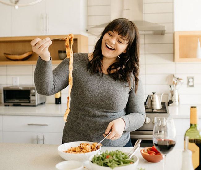 A person in a gray sweater joyfully holds a fork with a long strand of pasta above a bowl on a kitchen counter. There are salad greens, a glass of red wine, and various kitchen items visible in the background.