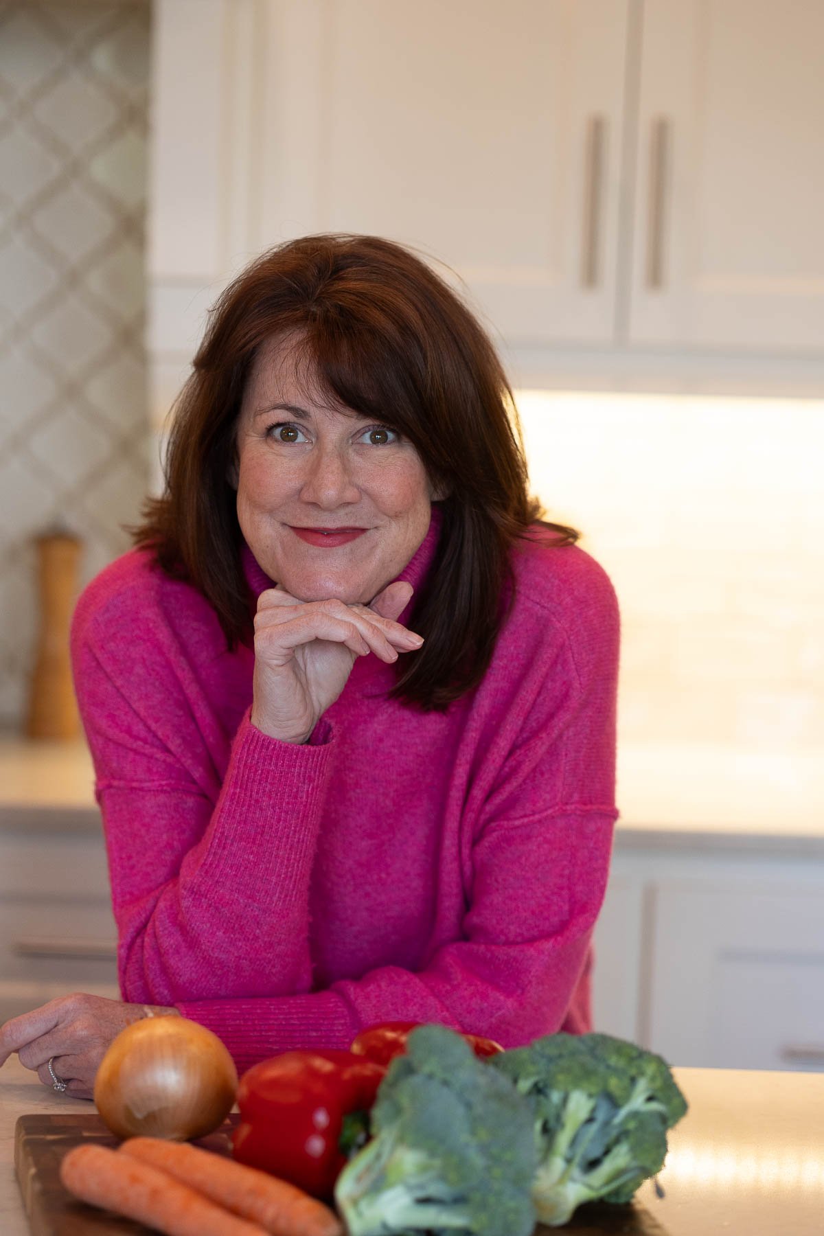 Woman in a pink sweater smiles, leaning on a kitchen counter with vegetables like broccoli, tomatoes, and carrots in the foreground. The kitchen has light-colored cabinets and a warm, cozy feel.