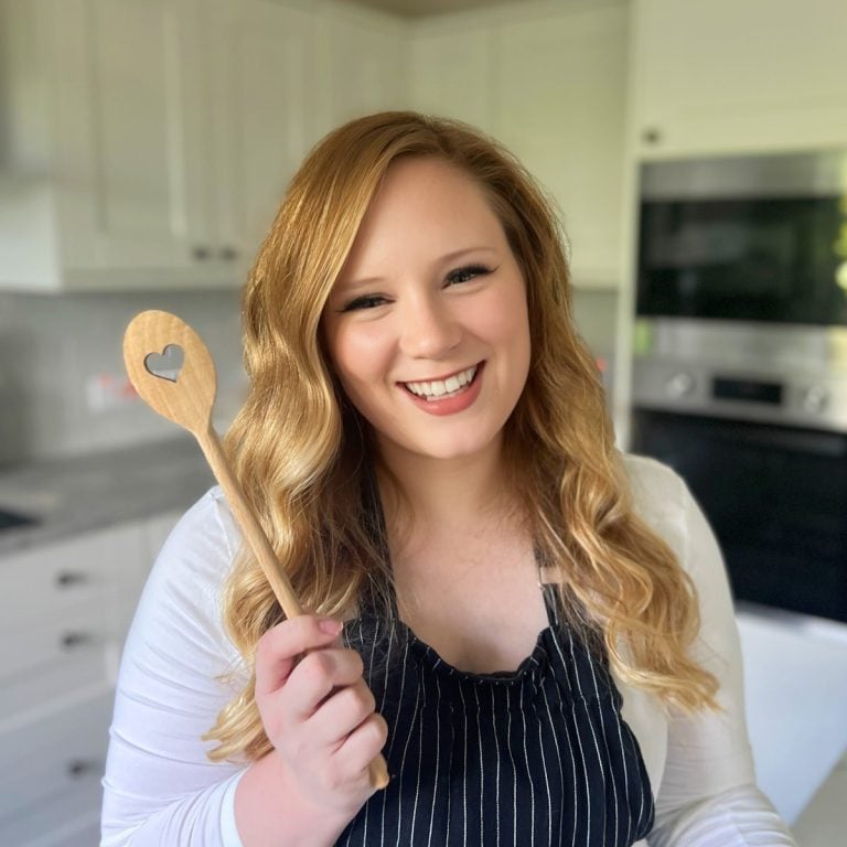 A woman with long wavy blonde hair smiles while holding a wooden spoon with a heart-shaped cutout. She is wearing a black apron over a white shirt and standing in a bright, modern kitchen.