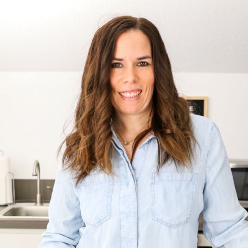 A woman with long, wavy brown hair is smiling and standing in a bright kitchen. She is wearing a light blue denim shirt. The background shows a sink, a microwave, and some kitchen appliances.