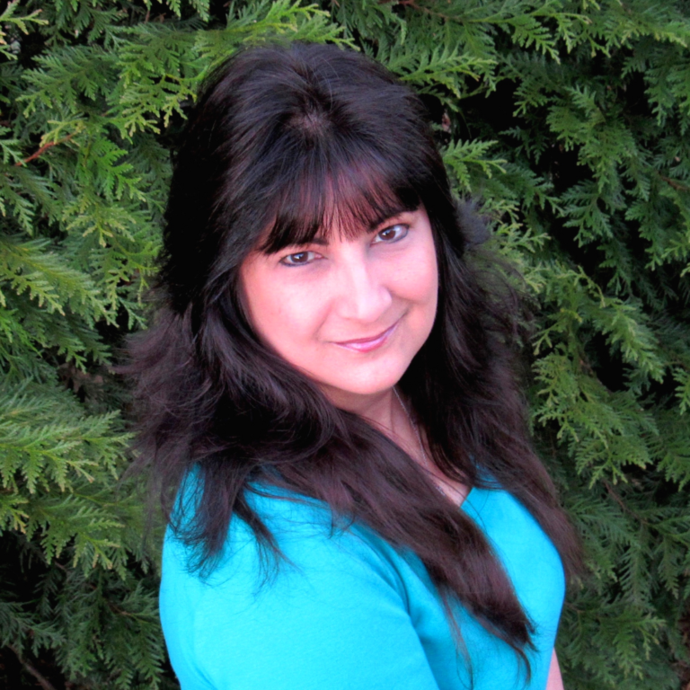 A woman with long, dark hair stands in front of a backdrop of lush green foliage. She is wearing a turquoise top and has a gentle smile on her face. Her head is slightly turned to the side, with her eyes looking directly at the camera.
