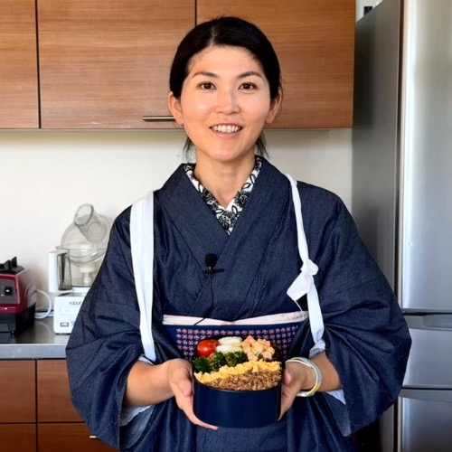 A person in a dark blue kimono smiles while holding a bento box filled with colorful food, including broccoli, tomatoes, and rice. They stand in a modern kitchen with wooden cabinets and silver appliances in the background.