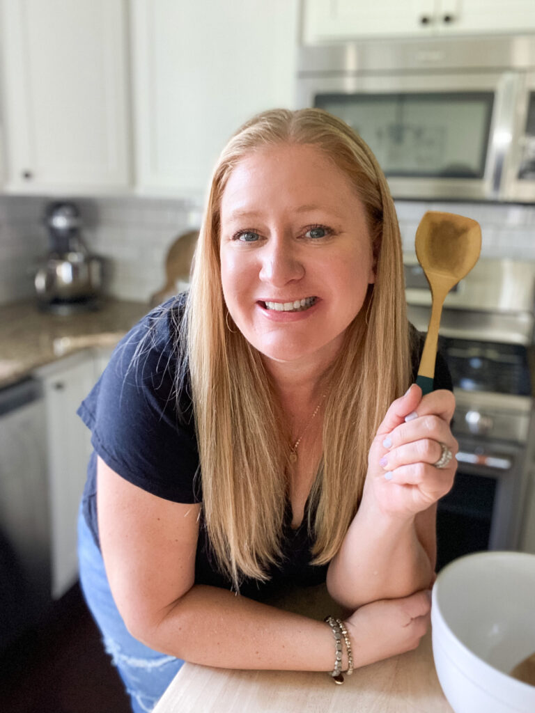 A woman with long blonde hair and wearing a black t-shirt smiles at the camera while holding a wooden spoon in a kitchen setting. The kitchen features white cabinets and stainless steel appliances.