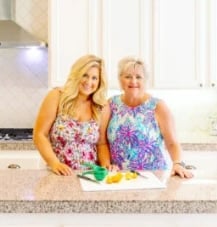 Two women smiling in a kitchen, standing behind a counter with chopped fruits and kitchen utensils. The kitchen has white cabinets and a stove in the background.