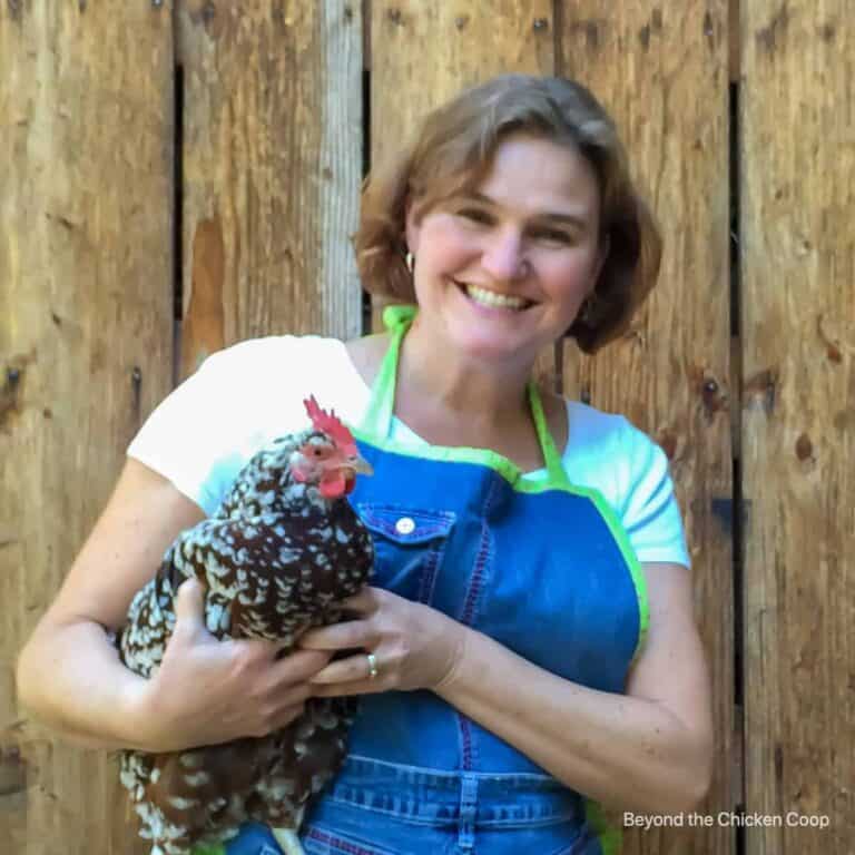 A smiling woman with short brown hair, wearing a white shirt and a blue apron, stands in front of a wooden wall while holding a speckled chicken in her arms.