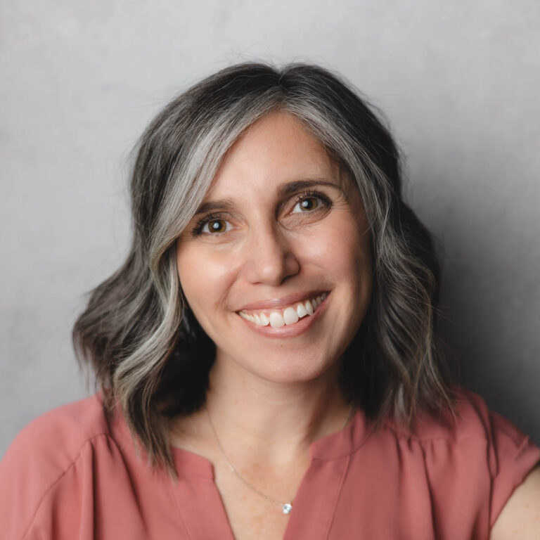 A woman with shoulder-length wavy hair with gray streaks smiles warmly at the camera. She is wearing a pink blouse and a delicate necklace. The background is a neutral gray.