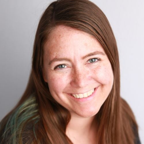 Smiling woman with long brown hair and blue eyes, wearing a dark top, against a plain background.