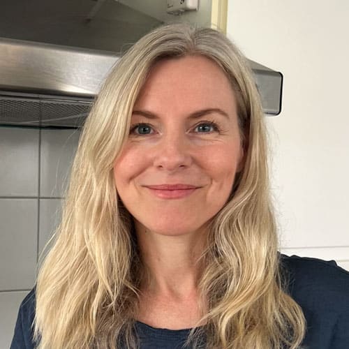 A woman with long, wavy, blond hair smiles softly. She is wearing a dark blue top and stands indoors in what appears to be a kitchen, with part of a stainless steel range hood and white tiled backsplash visible in the background.