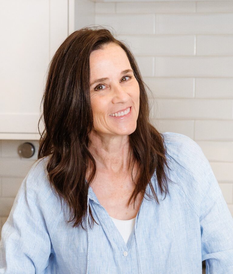 A person with long dark hair, wearing a light blue shirt over a white top, smiles while standing in a kitchen with white tiled walls in the background.