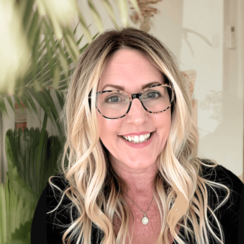 Smiling woman with long blonde hair and glasses, wearing a black top and a necklace with a pendant. She is standing in front of greenery, possibly indoor plants.