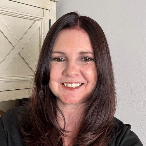 Smiling woman with long, dark brown hair wearing a black top, standing indoors near a light-colored cabinet with decorative paneling.