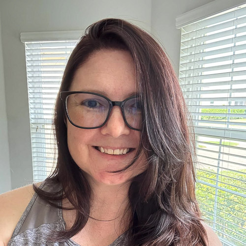 A person with long, dark brown hair and glasses smiles indoors. They are wearing a gray top, and sunlight filters through the window blinds behind them.