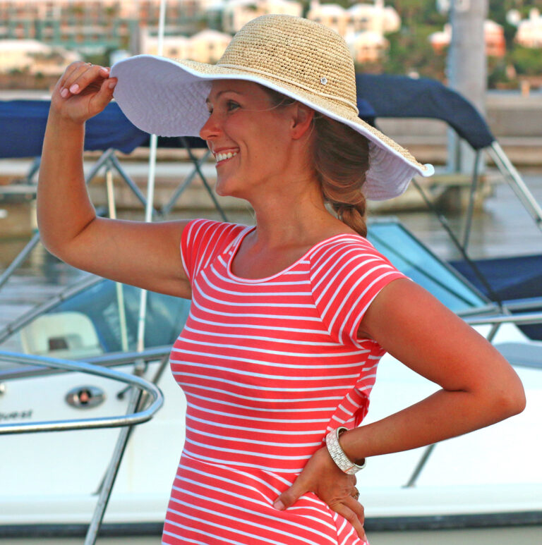 A woman wearing a large sun hat and a red and white striped dress is standing in front of docked boats. She is smiling, looking to the left, and raising her right hand to adjust her hat. The background features more boats and a blurry view of buildings.