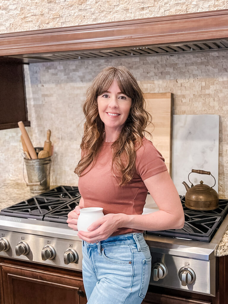A woman with long brown hair stands in a kitchen, holding a white mug. She is wearing a brown short-sleeved top and light blue jeans. Behind her is a stovetop with a kettle and rolling pins in a container. The backsplash features light-colored tiles.