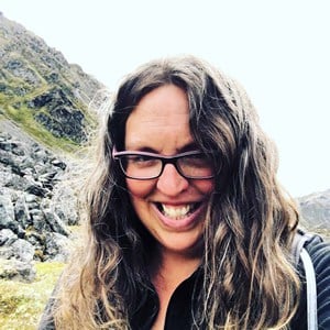 Woman with long, wavy hair and glasses smiling outdoors. She is wearing a black top and standing in front of a rocky, mountainous landscape. The sky is overcast.
