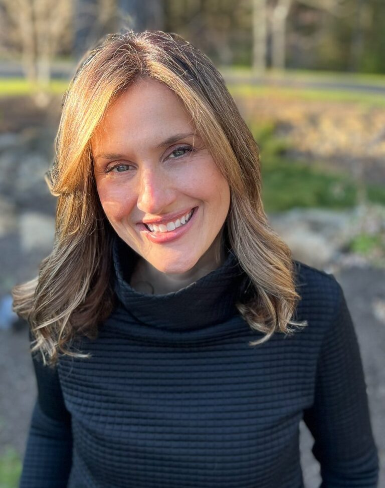 Smiling woman with shoulder-length, light brown hair, wearing a black textured top, stands outdoors. Sunlight highlights her hair against a blurred garden background.