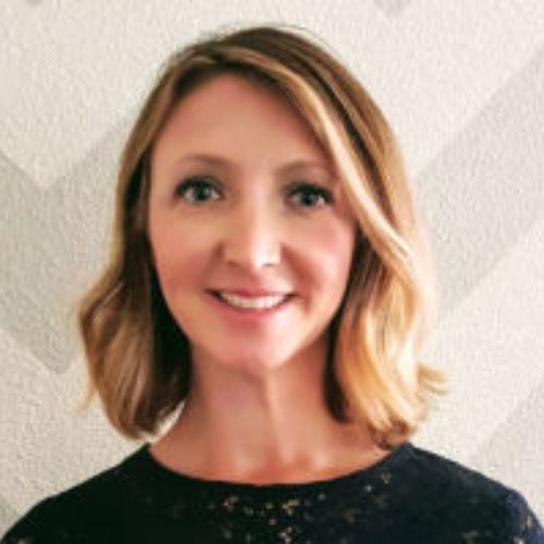 A woman with shoulder-length blonde hair and a side part smiles at the camera. She is wearing a dark top and is posed in front of a textured, light-colored wall with a subtle geometric pattern.