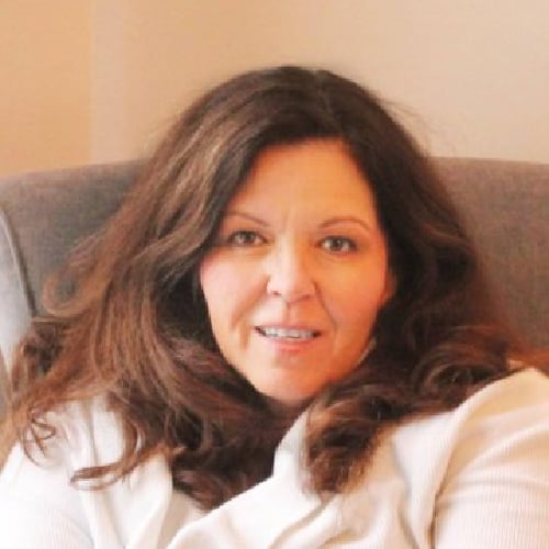 A person with long brown hair sitting on a gray chair, wearing a white top, and looking at the camera with a neutral expression.