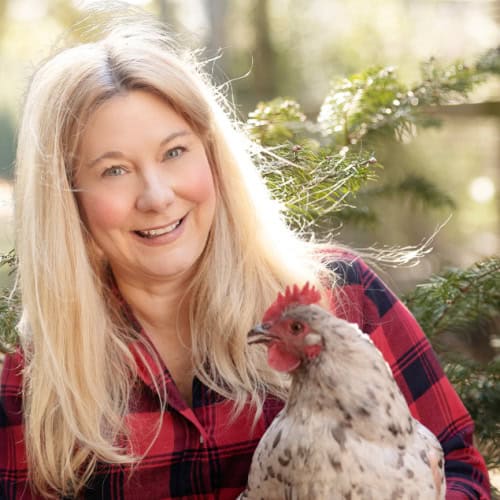 A woman with long blonde hair, wearing a red and black plaid shirt, smiles while holding a light-colored chicken with a red comb. The background features outdoor greenery, suggesting a natural setting.