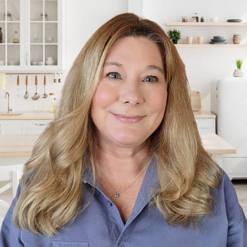 A smiling woman with long, blond hair is standing in a modern kitchen. She is wearing a blue shirt, and behind her are white cabinets, open shelves with decor, and a kitchen island. The space is well-lit and tastefully decorated.