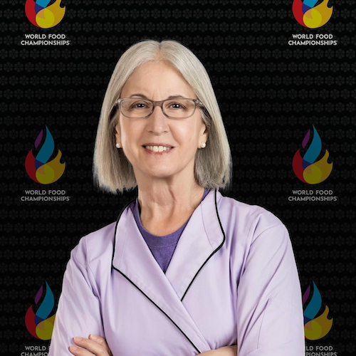 An older adult with gray hair and glasses smiles, arms crossed, wearing a light purple jacket. In the background is a World Food Championships logo pattern featuring flame-like shapes in blue, red, and yellow.