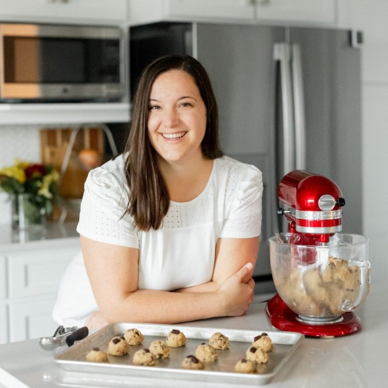 A woman in a white blouse smiles in a kitchen. She stands next to a baking tray with cookie dough pieces and a red stand mixer filled with more dough. Appliances and flowers are in the background.