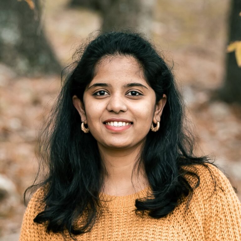 Smiling person with long black hair wearing a mustard sweater and hoop earrings, standing outdoors in a forested area with trees and fallen leaves in the background.