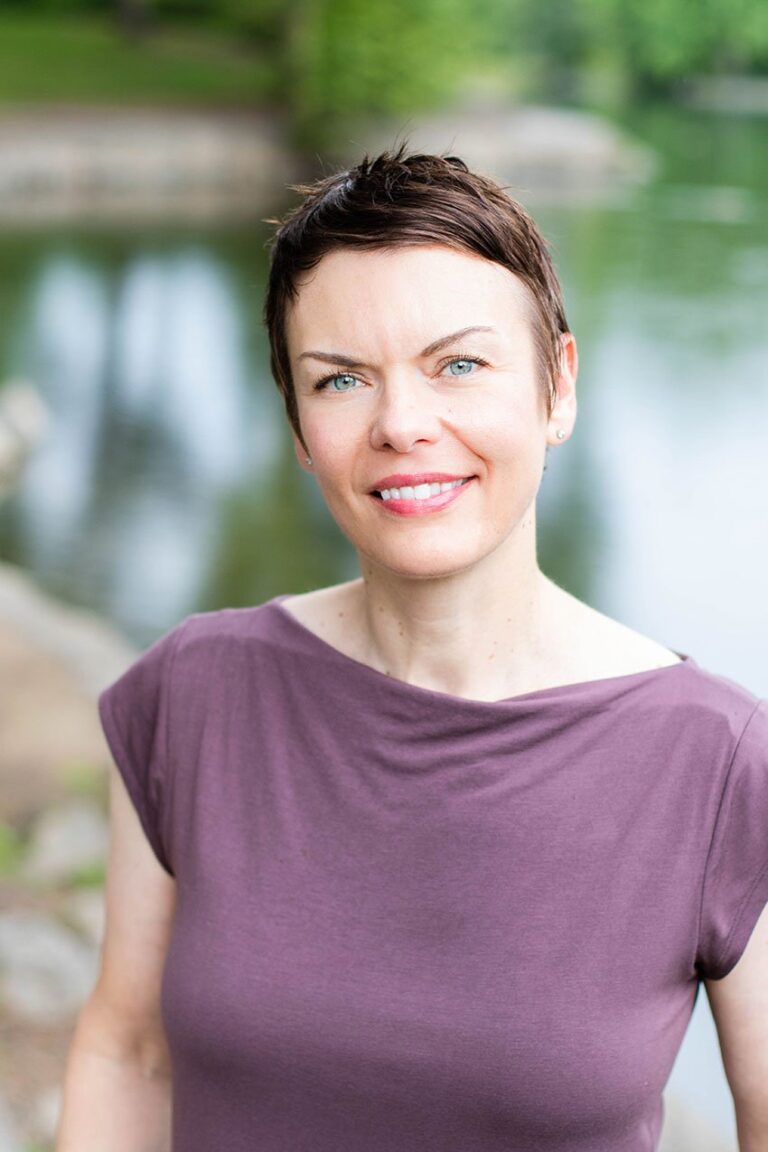 A person with short hair and a purple top smiles at the camera while standing outdoors near a body of water, with trees in the background.