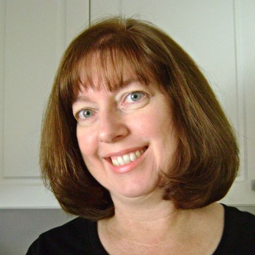 A woman with shoulder-length brown hair and bangs smiles at the camera. She is wearing a black top and is standing against a background of white cabinets.