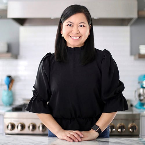 A woman with long dark hair, wearing a black blouse and a watch, stands smiling in a modern kitchen. The background features a stove, white subway tiles, and appliances, including a blue mixer.