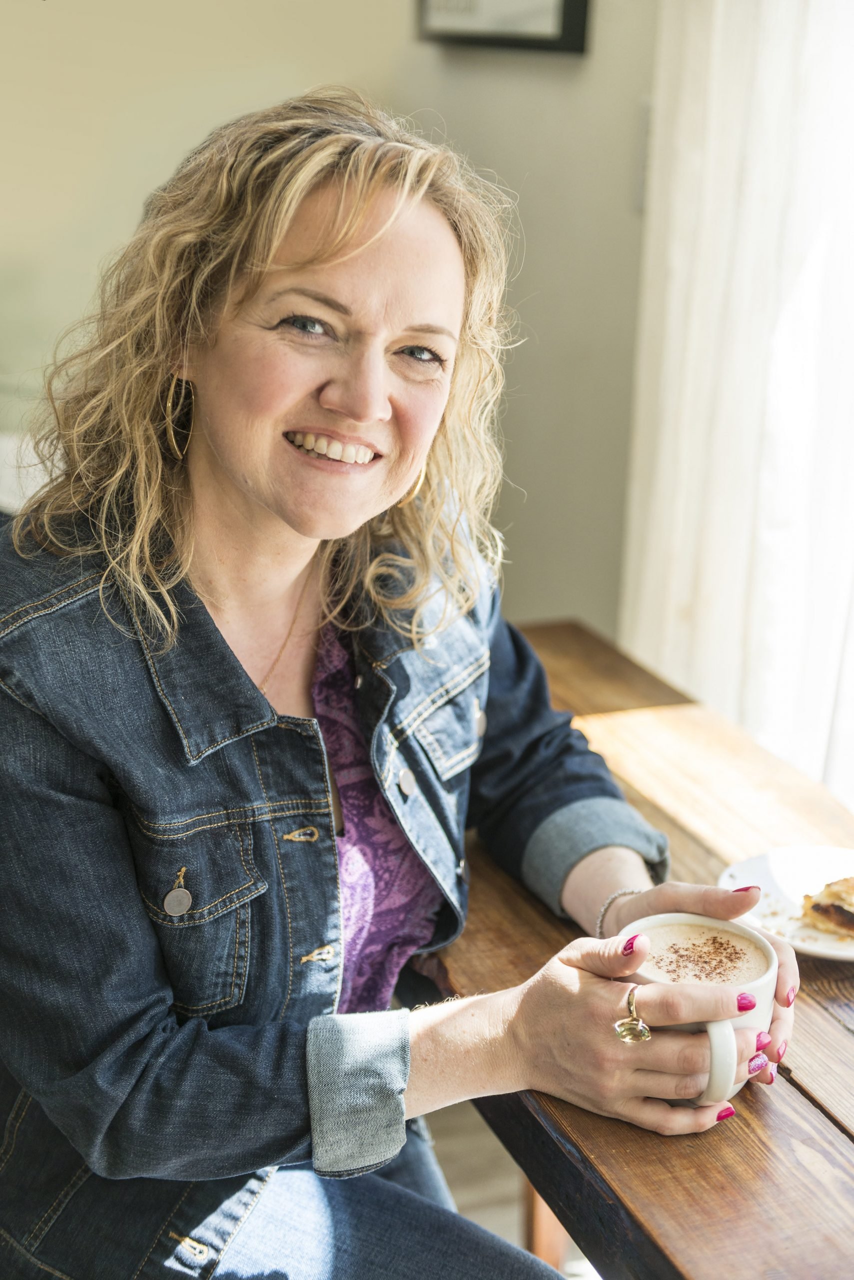 A smiling woman with curly blonde hair sits at a wooden table holding a mug of coffee. She is wearing a denim jacket over a purple top. Sunlight streams in from a window to the right, casting a warm glow on the scene.