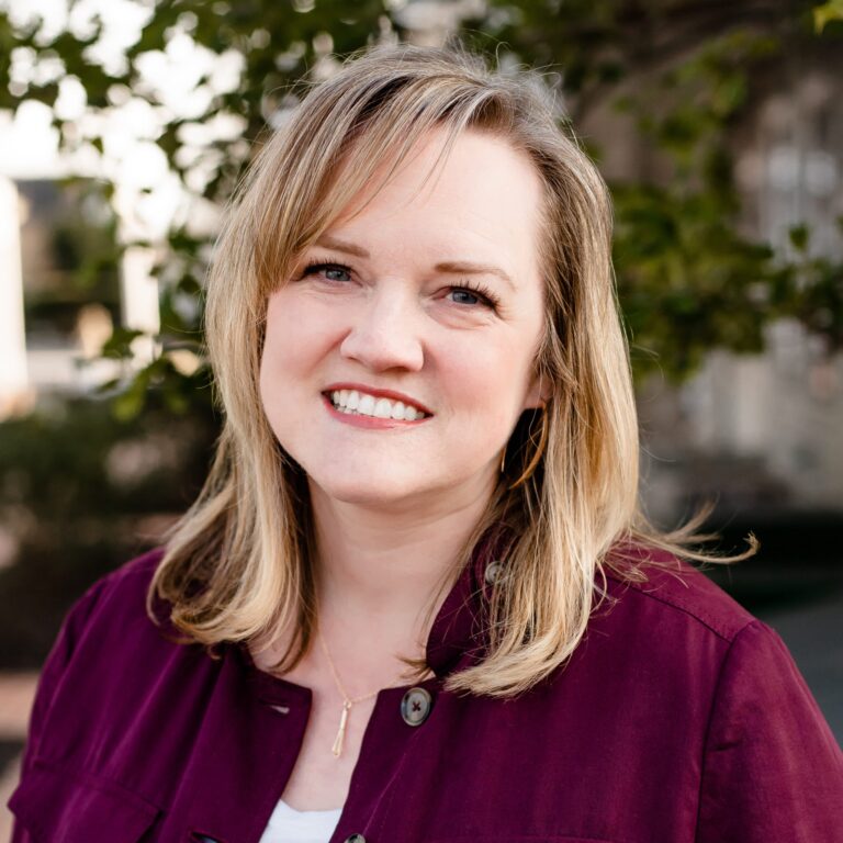 Smiling person with shoulder-length blonde hair wearing a maroon jacket and white top stands outdoors. Blurred greenery in the background.