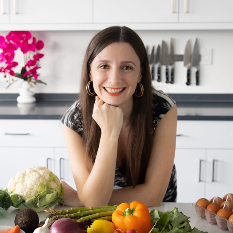 A woman with long brown hair and hoop earrings is smiling in a kitchen. She rests her chin on her hand among a variety of fresh vegetables, including cauliflower, bell pepper, and asparagus. In the background are white cabinets, a knife rack, and an orchid plant.