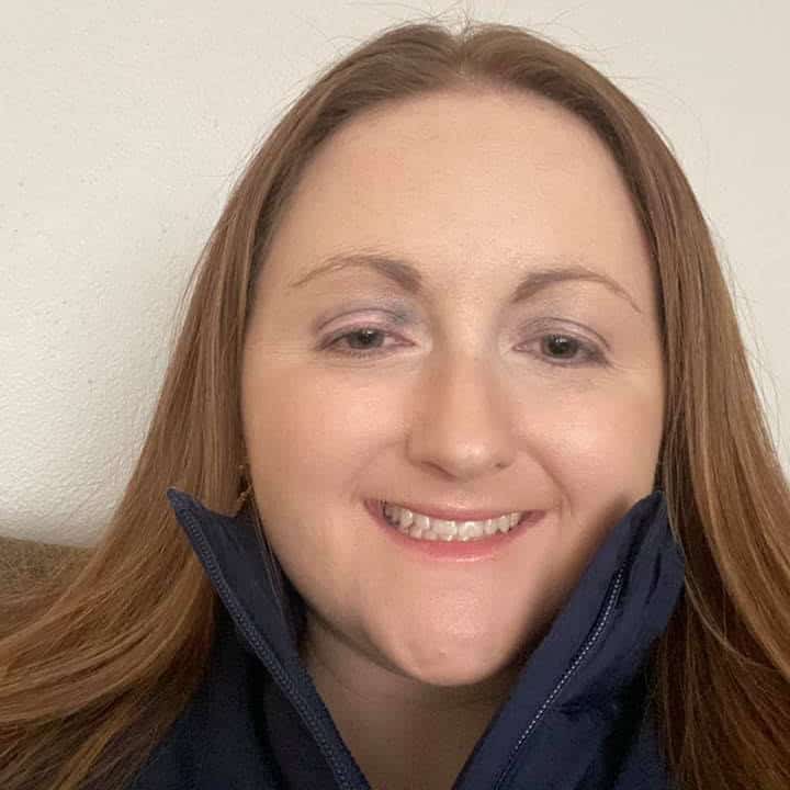 A woman with long, straight brown hair is smiling at the camera. She is wearing a navy blue jacket with the collar slightly raised. The background is plain white.