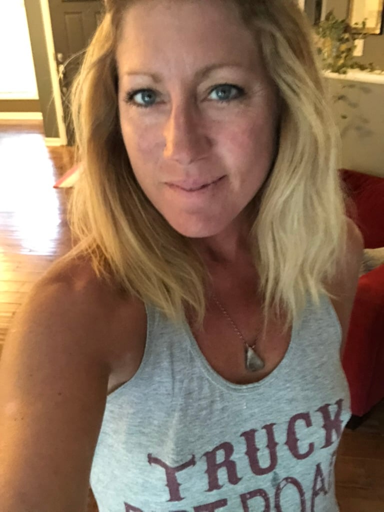 A woman with shoulder-length blonde hair and blue eyes smiles slightly at the camera. She wears a gray tank top and a necklace with a triangular pendant. The background shows a cozy, well-lit room with wooden floors.