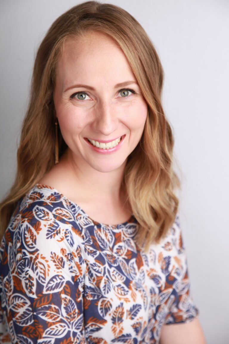 Smiling woman with wavy blonde hair wearing a patterned blue and orange top, posing against a plain light background.
