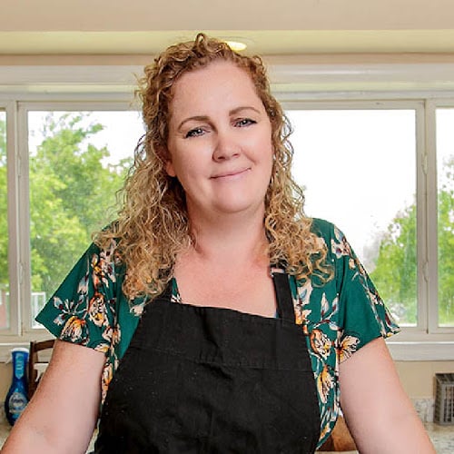 Woman with curly hair wearing a floral top and a black apron, standing in a kitchen with large windows showing greenery outside. She is smiling softly.