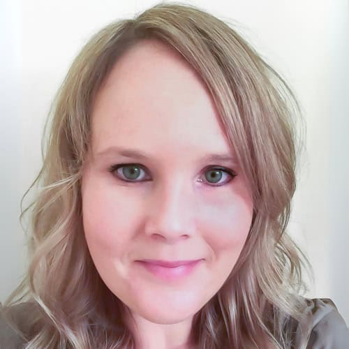 A person with light blond hair and green eyes is looking towards the camera with a neutral expression. They have slightly wavy hair that falls to their shoulders and are positioned against a plain, light-colored background.