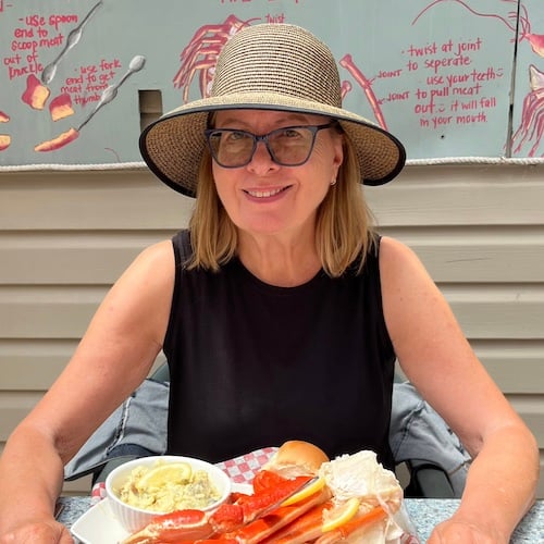 A woman wearing a wide-brimmed hat and glasses sits at a table with a plate of crab legs and a side dish. She is smiling and is against a backdrop with illustrated instructions for eating crab.
