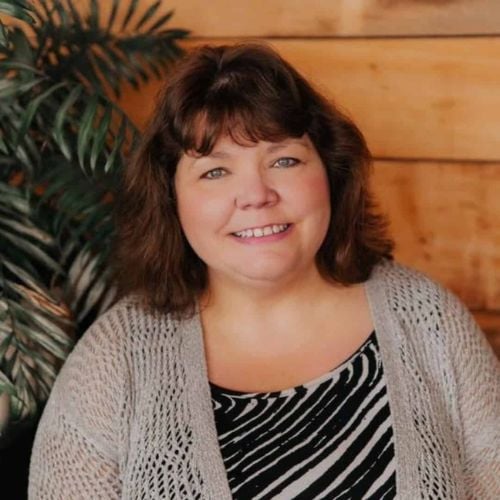 A woman with shoulder-length brown hair smiles while sitting in front of a wooden wall and a leafy plant. She is wearing a light gray cardigan over a zebra-patterned shirt.
