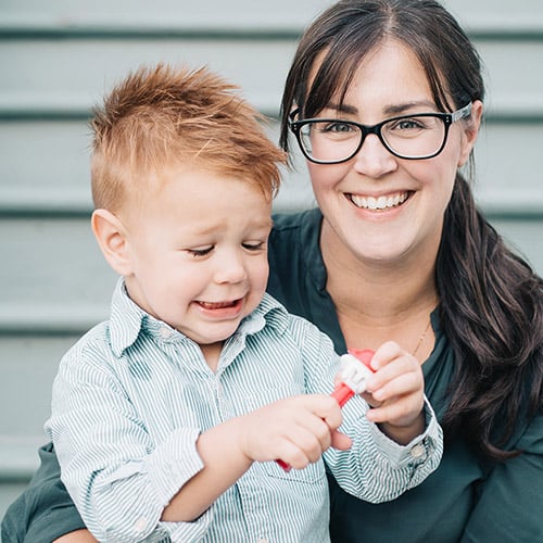A woman with glasses smiling while holding a young boy with spiky hair. The boy, wearing a striped shirt, looks at a toy in his hands with a slight grin. They are sitting on steps.