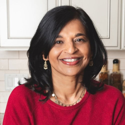 A woman with shoulder-length black hair, wearing a red sweater and gold earrings, smiles at the camera. The background shows a white kitchen with a blue-gray backsplash, wooden utensils, and bottles neatly placed on the countertop.