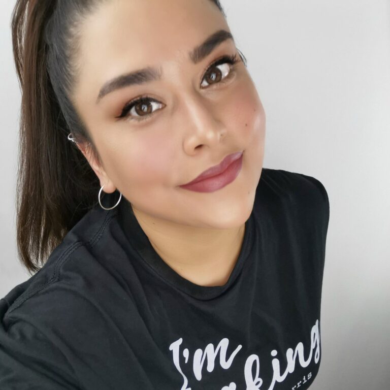 A person with long dark hair tied back, wearing a black shirt with white text, smiles at the camera. They have hoop earrings and a natural makeup look. The background is plain white.