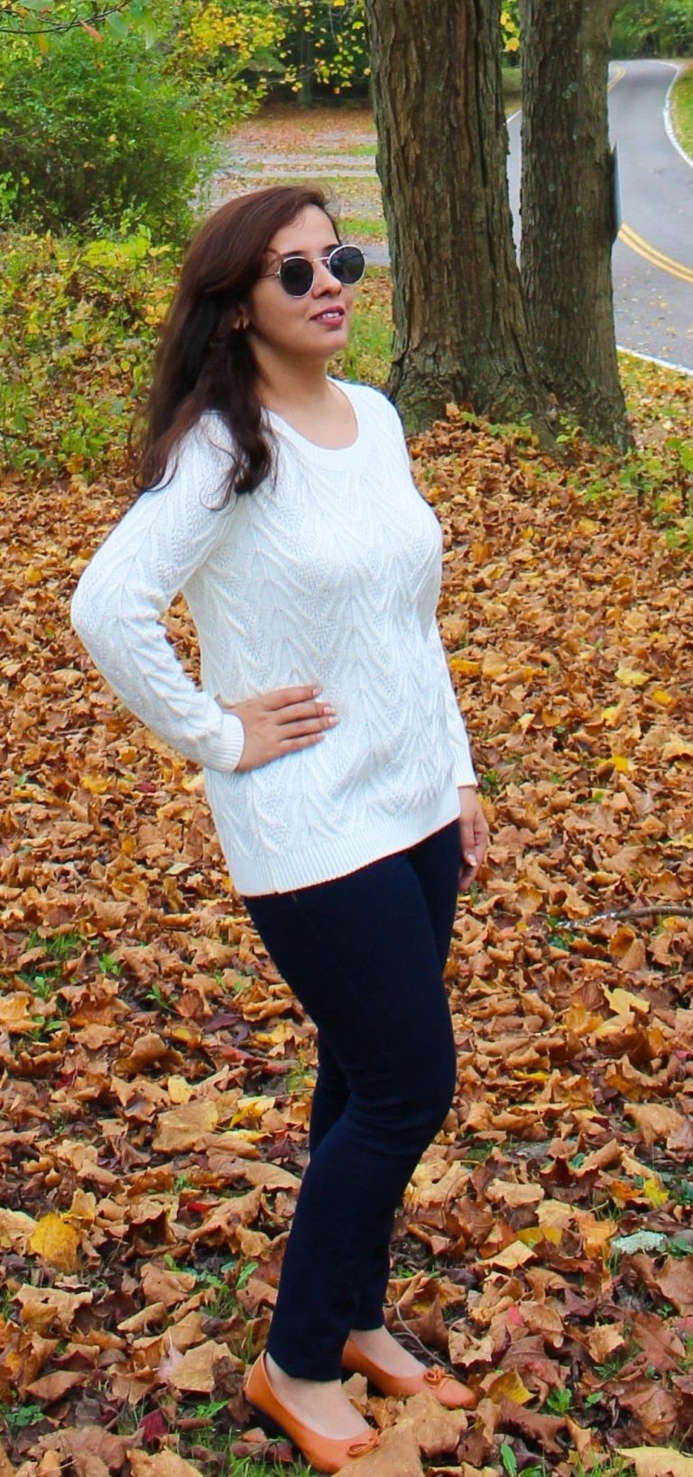 A woman wearing a white sweater, dark pants, and sunglasses stands on fallen autumn leaves near a road, posing with one hand on her hip and trees in the background.