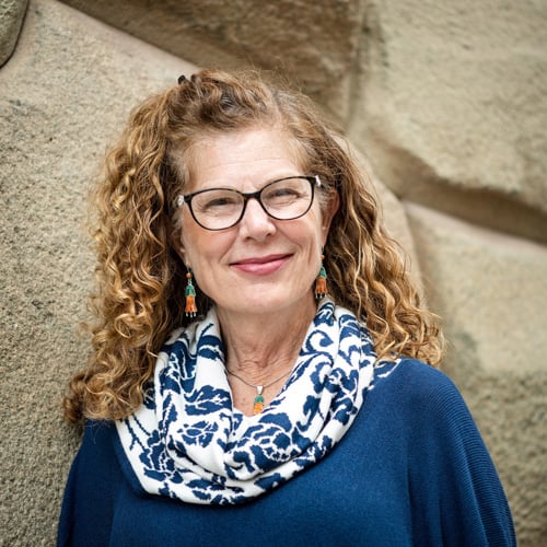 A woman with curly hair and glasses smiles while standing in front of a textured stone wall. She is wearing a blue outfit with a patterned scarf and colorful earrings.