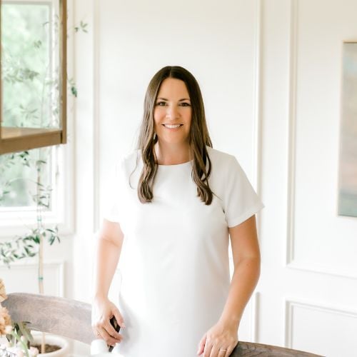 A woman with long brown hair, wearing a white dress, stands in a bright room beside a wooden chair. She is smiling, and there is a window with greenery to her left. The room has white walls and is warmly lit.