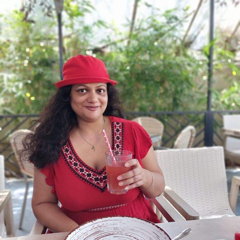 A person in a red dress and matching hat sits at an outdoor cafe, holding a red drink and smiling. The background features green plants and white chairs.