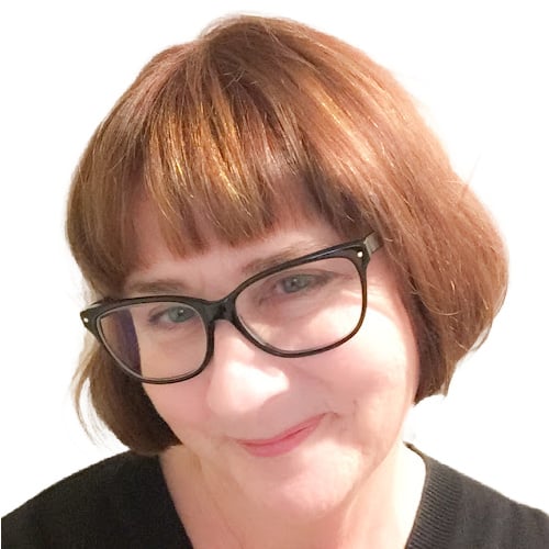 A person with short auburn hair and glasses is smiling softly. They are wearing a black top and have a white background.